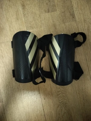 Svarta benskydd med gula ränder - Säljer ett par svarta benskydd med vita diagonala ränder och justerbara kardborreband. Snygg sportig look och perfekt för dig som lirar fotboll. Tillverkade i slitstark plast och syntetmaterial för bra skydd och komfort.