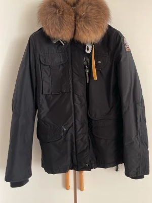Svart Parajumpers parkajacka päls - Säljer min svarta Parajumpers parkajacka med stor fluffig päls runt kragen och pälsfoder på insidan. Jackan har flera stora fickor, gul detalj vid dragkedjan och patch på ärmen. Perfekt för kalla dagar och riktigt snygg streetstil.