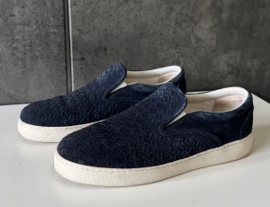 Blå slip-on sneakers från Bottega Veneta - Snygga slip-on sneakers från Bottega Veneta i mörkblå mocka med det klassiska flätade mönstret. Skorna har en vit platt gummisula och rund tå. Insidan är ljus och mjuk för extra komfort. Perfekta för dig som gillar stilrena och lyxiga detaljer.