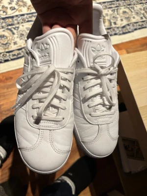 Vita Adidas Gazelle sneakers i skinn - Säljer ett par fräscha Adidas Gazelle sneakers i helvitt skinn med klassiska tre ränder på sidan och guldfärgad Gazelle-text. Skorna har snörning, rund tå och platt sula. Perfekta för dig som gillar stilrena och cleana sneakers.