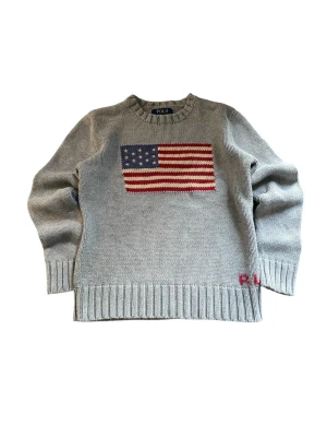Grå stickad tröja Polo Ralph Lauren - Grå stickad tröja från Polo Ralph Lauren med amerikansk flagga framtill och broderad RL-detalj i rött vid nederkanten. Tröjan har rund halsringning, ribbade muddar och är långärmad. Perfekt för dig som gillar klassisk och stilren design.