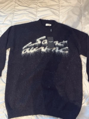 Svart stickad tröja Saint Laurent - Cool svart stickad tröja från Saint Laurent med vit fuzzy logga framtill. Tröjan har rund hals och långa ärmar, perfekt för dig som gillar exklusiva märken och vill ha en unik look. Materialet är mjukt och ger en lyxig känsla.