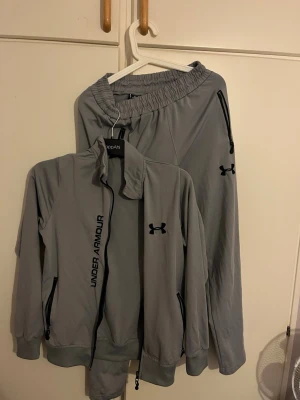 Under amour - Bilderna visar ett grått träningsset från Under Armour med jacka och byxor, båda med svarta dragkedjor och logotyp. Ingen sko syns på bilderna.