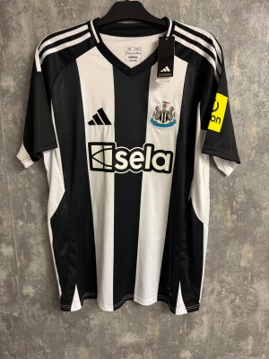 Newcastle ISAK - ny storlek 2XL - Snygga fotbollsskor från Adidas i svart och vitt med klassiska ränder och Newcastle United-logga. Skorna har randigt mönster, platt sula och är tillverkade i syntetmaterial. Perfekta för dig som älskar fotboll och vill sticka ut på planen.