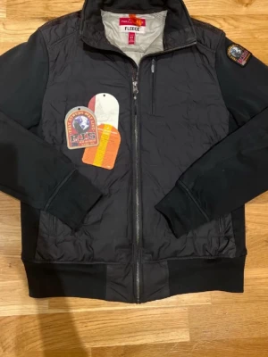 Svart bomberjacka från Parajumpers - Svart bomberjacka från Parajumpers med quiltad framsida, ribbade muddar och dragkedja. Jackan har en patch på ärmen och en säkerhetsficka på insidan. Materialet är polyester och fleece, perfekt för dig som vill ha en snygg och funktionell jacka.