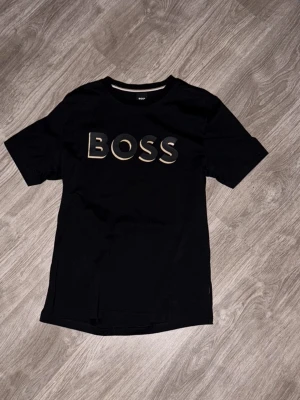 BOSS T-shirt - Svart t-shirt från BOSS med stort svart-guldfärgat BOSS-tryck på bröstet. Perfekt för dig som gillar stilrena och trendiga plagg.