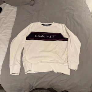 Vit sweatshirt från Gant med tryck - Vit sweatshirt från Gant med ett mörkblått band och röd linje över bröstet där Gant-loggan är tryckt. Tröjan har rund hals, ribbade muddar och är långärmad. Perfekt för dig som gillar klassisk och sportig stil. I storlek 11-12 år. Går att diskutera pris!