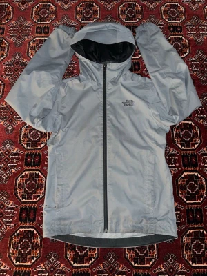 Ljusgrå vindjacka från The North Face - Säljer en ljusgrå vindjacka från The North Face med huva och hel dragkedja framtill. Jackan har en stilren design och är tillverkad i slitstark polyester. Perfekt för blåsiga dagar och outdoor-aktiviteter. Loggan är tryckt på bröstet.