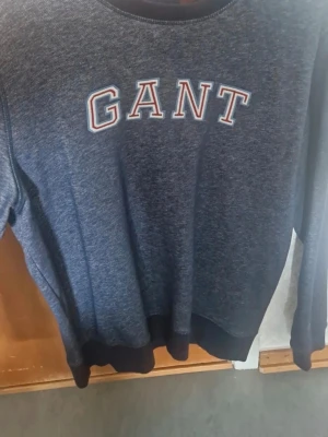 Mörkblå sweatshirt från GANT - Säljer en mörkblå sweatshirt från GANT i storlek L. Tröjan har klassisk rund hals och stora vita bokstäver med röd kant på bröstet. Muddar vid ärmslut och nederkant ger en skön passform. Perfekt för chill dagar och enkel att matcha med jeans.