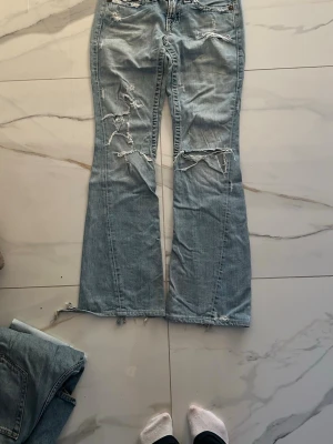 True Religion ljusblå bootcut jeans - Säljer ett par ljusblå bootcut jeans från True Religion med slitna detaljer och grova sömmar. Jeansen har låg midja, klassiska fem fickor och snygga knappar med logga. 