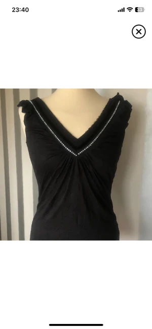 Svart topp med pärldetaljer - Snygg svart topp med v-ringning och pärldetaljer längs halsen. Toppen har en lätt rynkad effekt framtill och är ärmlös med små volangdetaljer vid axlarna. Materialet känns mjukt och har en diskret struktur. Perfekt för dig som gillar stilrena detaljer.