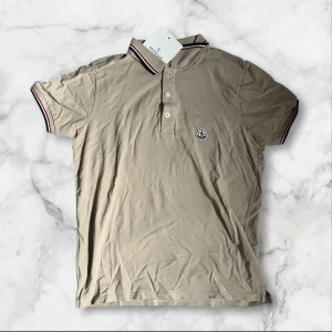 Beige piké från Moncler - Säljer en beige piké från Moncler med klassisk krage och korta ärmar. Pikén har knappar framtill och Moncler-logga broderad på bröstet. Kragen och ärmsluten har snygga ränder i rött, vitt och blått. Perfekt för en stilren och avslappnad look.