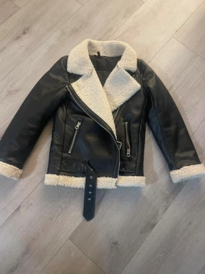 Svart teddyfodrad skinnjacka - Säljer en svart skinnjacka med krämvit teddyfoder på krage, ärmslut och nederkant. Jackan har två snedställda dragkedjefickor, bälte i midjan och grov dragkedja framtill. Perfekt för dig som vill ha en cool och varm look i höst och vinter.