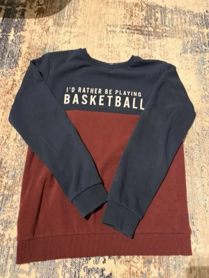 Blå och vinröd sweatshirt från Name It - Säljer en cool sweatshirt från Name It med texten 'I’d rather be playing basketball' på bröstet. Tröjan är tvåfärgad med marinblå upptill och vinröd nedtill, har långa ärmar och ribbade muddar. Perfekt för dig som gillar basket och vill ha en avslappnad stil.