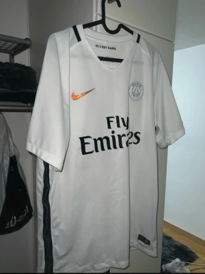 Vit PSG fotbollströja från Nike - Säljer en vit Paris Saint-Germain fotbollströja från Nike med svart Fly Emirates-tryck och orange Nike-logga på bröstet. Tröjan har svarta ränder längs sidorna och PSG-emblem på vänster bröst. Perfekt för dig som älskar fotboll och PSG.