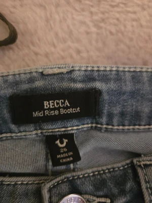 True Religion BECCA Bootcut Jeans - Säljer ett par blå bootcut jeans från True Religion, modell BECCA Mid Rise Bootcut. Jeansen har klassisk femficksdesign, snygga detaljer på bakfickorna och silverfärgade knappar. Materialet är jeans med en tvättad look och midwaist passform.