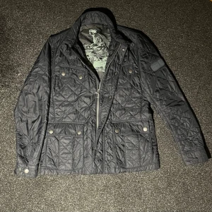 Svart quiltad jacka från Barbour - Quiltad marinblå jacka från Barbour International med snygga tryckknappar och dragkedja framtill. Jackan har flera fickor och ett coolt tryck på insidan. Perfekt för dig som gillar stilren design med lite edge.