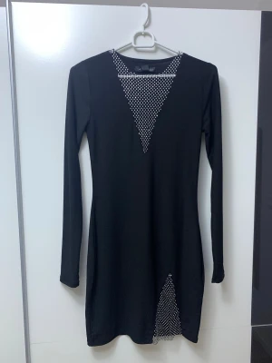 Svart klänning med glittriga detaljer - Svart kortklänning med lång ärm och slim passform. Klänningen har glittriga meshdetaljer i silver vid halsringningen och på sidan av nederdelen, vilket ger en snygg och unik look. Materialet är mjukt och stretchigt, perfekt för att sticka ut. Passar dig som gillar att glänsa.