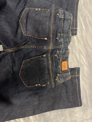 Mörkblå jeans Q3000 Samantha, storlek 27 - Snygga mörkblå jeans från Q3000 Samantha med kontrastsömmar och dubbla knappar i midjan. Klassisk femficksdesign och breda bälteshällor ger en cool look. Passar dig som gillar jeans med detaljer och markerad midja, är dessutom lågmidjade. Aldrig använda då dom inte passar