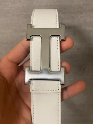 Vit och svart skärp med H-spänne - Snyggt och stilrent skärp i vitt och svart med ett stort H-format metallspänne i silverfärg. Skärpet är tillverkat i präglat skinn och har tydliga sömmar längs kanterna. Perfekt accessoar för att lyfta din outfit.