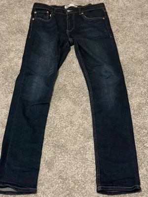Levi's 512 mörkblå jeans - Säljer ett par Levi's 512 jeans i mörkblå tvätt med klassisk femficksdesign och kontrastsömmar. Modellen har en slim passform och är tillverkad i en mjuk blandning av bomull, polyester, viskos och elastan för extra komfort och stretch. Perfekt för dig som gillar stilrena jeans.