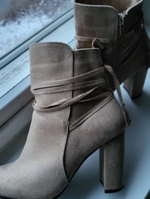Beige mocka boots med klack - Snygga beige boots i mjuk mocka med hög klack och rund tå. Skorna har dekorativa snören runt skaftet och dragkedja på sidan för enkel på- och avtagning. Perfekt för dig som gillar stilrena detaljer och vill ha ett par boots som sticker ut.