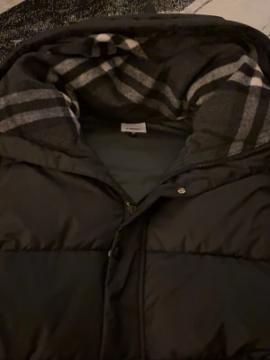 Svart pufferjacka från Burberry - Svart pufferjacka från Burberry med huva och klassiskt rutigt foder. Jackan har dragkedja och knappar framtill, två sidofickor med dragkedja och snygga detaljer på knapparna. Perfekt för kalla dagar och har en loose passform.