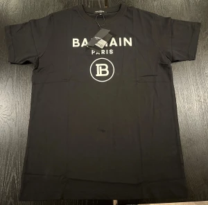 Svart Balmain Paris t-shirt - Svart t-shirt från Balmain Paris med vit logotyp och tryck framtill. Klassisk rund hals och korta ärmar. Tillverkad i mjuk bomull som känns skön mot huden. Perfekt för dig som gillar stilrena och lyxiga plagg med tydlig branding.