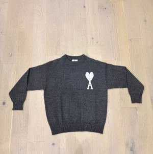 Mörkgrå stickad tröja från Ami Paris - Säljer en mörkgrå stickad tröja från Ami Paris med vit hjärtformad A-logga på bröstet. Tröjan har rund hals, ribbade muddar och är långärmad. Perfekt för dig som gillar stilrena och snygga detaljer.