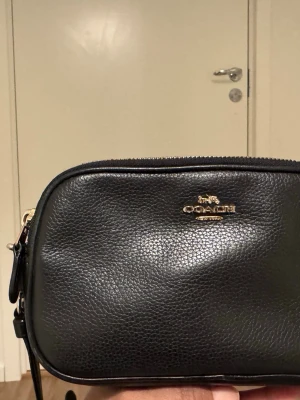 Svart axelväska från Coach i skinn - Snygg svart axelväska från Coach i äkta skinn med guldfärgade detaljer och dragkedja. Väskan har en stilren, rektangulär form och justerbar axelrem. Perfekt storlek för det viktigaste och har ett fack på insidan. Coach-logga i guld på framsidan.