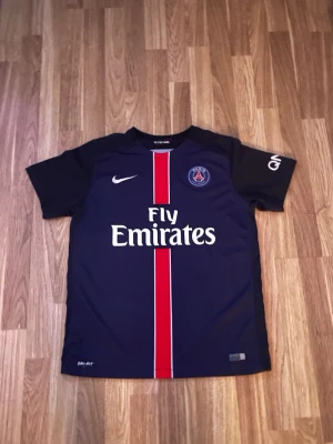 PSG matchtröja – Nike – Fly Emirates - Säljer en stilren Paris Saint-Germain matchtröja från Nike. Klassisk mörkblå design med den ikoniska röda/vita mittlinjen, PSG-emblem på bröstet och Fly Emirates som sponsor.  Tröjan är i fint skick utan hål eller fläckar. Perfekt för matchdag, träning eller som streetwear. Aldrig använd 