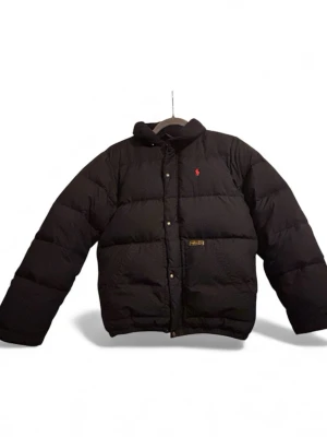 Svart pufferjacka från Polo Ralph Lauren - Säljer en svart pufferjacka från Polo Ralph Lauren med klassisk röd logga på bröstet och Polo RL & Co-märke på ena sidan. Jackan har hög krage, dragkedja och tryckknappar framtill. Perfekt för kalla dagar och riktigt snygg till streetwear-stilen.