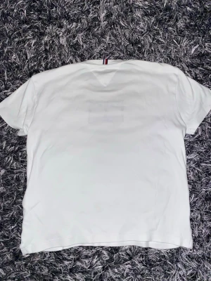 Vit t-shirt Tommy Hilfiger 152 - Vit t-shirt från Tommy Hilfiger med tryckt logga och text på bröstet. Rund hals och korta ärmar. Materialet är mjuk bomull och har en klassisk passform. Snygg detalj med Hilfiger-band i nacken. Perfekt för dig som gillar stilrena märkesplagg.