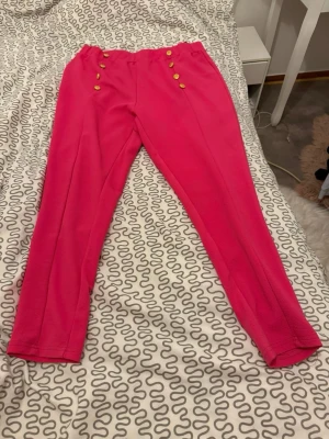 Rosa byxor med guldknappar - Säljer ett par snygga rosa byxor med hög midja och dekorativa guldknappar framtill. Byxorna har en rak passform och är gjorda i ett mjukt tyg som känns skönt mot huden. Perfekt för dig som vill sticka ut med färg och coola detaljer.