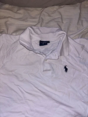 Vit Polo Sport pikétröja - Säljer en klassisk vit pikétröja från Polo Sport med korta ärmar och krage. Tröjan har två knappar vid halsen och den ikoniska Polo-loggan broderad i marinblått på bröstet. Perfekt för en clean och sportig look.