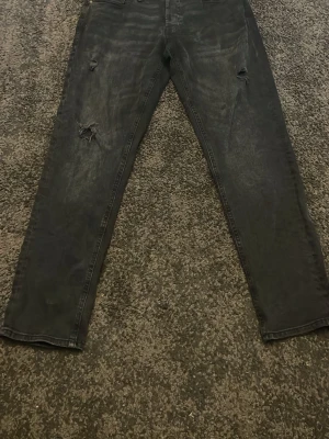 Jack and jones jeans - Säljer ett par svarta jeans med coola slitningar på benen. Jeansen har klassisk femficksdesign och raka ben. Materialet är jeans och färgen är mörk svart med lite tvättad look. Perfekt för dig som gillar en avslappnad och trendig stil.