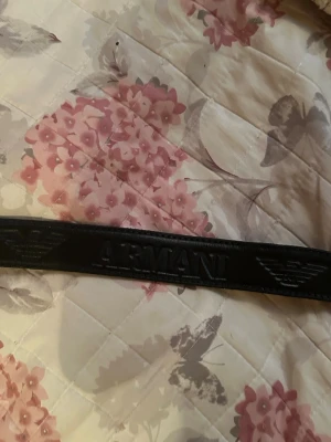 Svart skärp från Armani i skinn - Snyggt svart skärp från Armani med präglad logga och silverfärgat spänne. Skärpet är gjort i skinn och har en stilren design med metallplatta där märket syns tydligt. Perfekt för att lyfta din outfit med en exklusiv touch. Vet inte storleken tyvärr 