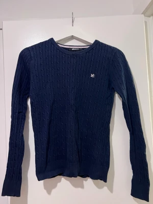 Mörkblå stickad tröja  - Snygg mörkblå stickad tröja från grina tricot med kabelstickat mönster och rund hals. Tröjan har lång ärm och en liten broderad logga på bröstet. Perfekt för dig som gillar klassisk och stilren design.