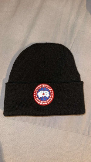 Svart Canada Goose mössa - Svart mössa från Canada Goose med klassisk rund logotyp framtill i rött, vitt och blått. Mössan är ribbstickad och har uppvikt kant. Materialet är mjukt och stickat, perfekt för kalla dagar. Snygg och enkel design som passar till det mesta. Jag har använd den knappt 