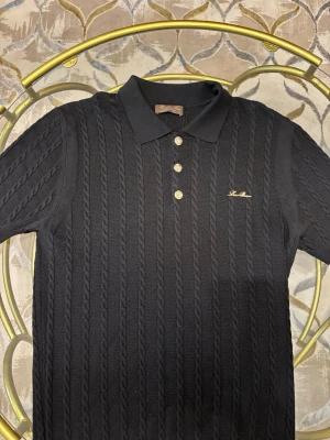 Svart pikétröja Loro Piana - Säljer en svart kabelstickad pikétröja från Loro Piana, tillverkad i Italien. Tröjan har klassisk krage, fyra guldknappar framtill och diskret broderad logga på bröstet. Materialet är 100% bomull och modellen har korta ärmar och en elegant struktur.