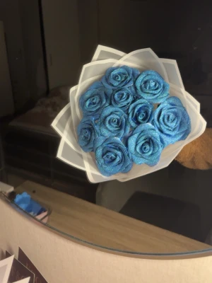 Blå glittrig ros-bukett - Bukett med konstgjorda rosor i intensivt blå färg och glittrigt ytskikt. Rosorna är tätt samlade och omslutna av ett stilrent vitt plastpapper med geometrisk form. Perfekt som dekor eller present till någon speciell.