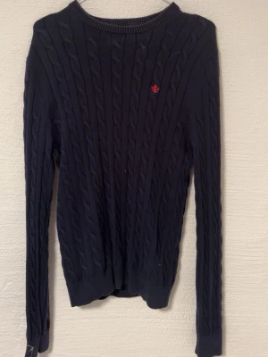 Mörkblå stickad tröja Morris M - Snygg mörkblå stickad tröja från Morris i storlek M. Tröjan har ett klassiskt kabelstickat mönster och en liten röd logga broderad på bröstet. Rund hals och långa ärmar, perfekt för dig som gillar stilrena och tidlösa plagg.