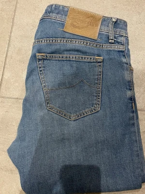 Ljusblå jeans från Jacob Cohen - Säljer ett par ljusblå jeans från Jacob Cohen med klassisk femficksdesign och snygga kontrastsömmar. Jeansen har rak passform och är tillverkade i mjukt bomullsmaterial. Perfekta för dig som gillar stilrena och tidlösa jeans. Jeans har tvyärr en liten defekt vid  skrevet men som är reparerat