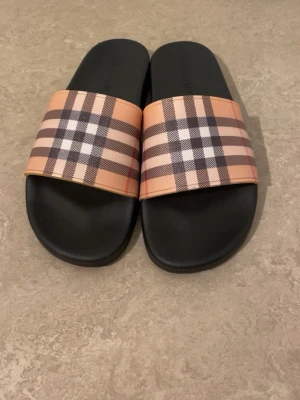 Burberry sandaler med klassiskt rutmönster - Säljer ett par snygga Burberry sandaler med bred rem i klassiskt beige, svart och rött rutmönster. Sulan är svart och platt, och remmen är tillverkad i syntetmaterial. Perfekt för sommaren och chill dagar. Ikonisk design som sticker ut.