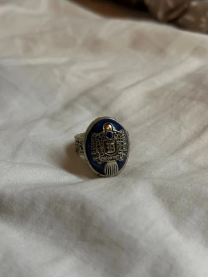 The vampire diaries - Damon's daylight ring - The vampire diaries merch - Damon's daylight ring. Säljer en cool ring i silverfärg med blått emalj och detaljerat emblem framtill. Ringen har en oval form och dekorerad kant, samt en liten blå sten upptill. Sidorna är prydda med mönster som ger en unik vibe. Perfekt för dig som gillar statement-smycken.
