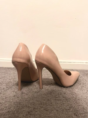 Beige pumps med spetsig tå och hög klack - Säljer ett par stilrena beige pumps med spetsig tå och hög klack. Skorna har en glansig finish och är tillverkade i syntetmaterial. Perfekta för dig som gillar klassisk och elegant stil. Klacken är smal och ger ett snyggt lyft.