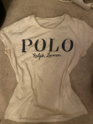 Vintage Vit t-shirt från Polo Ralph Lauren - Säljer en vit t-shirt från Polo Ralph Lauren i storlek S. T-shirten har korta ärmar och en rund halsringning. Stor, mörkblå logotyp med texten 'POLO Ralph Lauren' på bröstet. Skön och klassisk stil, perfekt till jeans eller shorts.