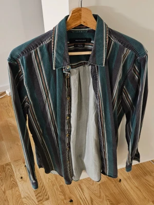 Randig skjorta från Urban Outfitters - Snygg skjorta med vertikala ränder i grönt, grått, beige och orange från Urban Outfitters. Klassisk krage, knappar framtill och långärmad. Skjortan är gjord i mjuk bomull och har en chill, loose passform som funkar till många olika stilar.