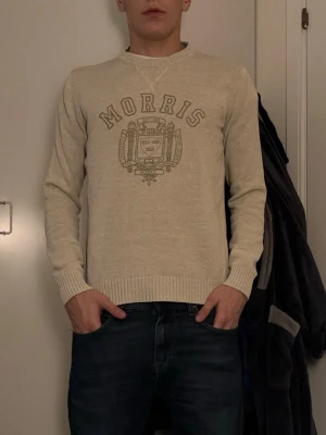Beige stickad tröja från Morris - Snygg beige stickad tröja från Morris Co i storlek medium. Tröjan har rund hals, ribbade muddar och ett stort Morris-tryck med emblem på bröstet. Passformen är normal och den är långärmad, perfekt för att hålla värmen under kyliga dagar. Modellen i bilden är 180 och väger 72 kg.