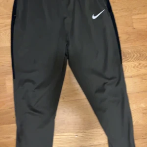 Mörkgröna sköna  träningsbyxor och tröja från Nike - Snygga mörkgröna träningsbyxor och tröja som en tracksuit från Nike med smal passform och elastisk midja. Byxorna har dragkedja vid fickorna och den klassiska Nike-loggan på framsidan. Perfekta för gymmet eller sportaktiviteter, tillverkade i lätt syntetmaterial. Pris kan diskuteras